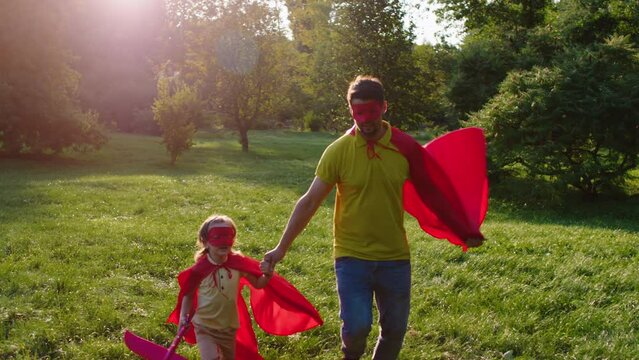 In front of the camera in the middle of the park in a sunny day superhero daddy and his cute small son wearing the red superhero suit and playing funny together