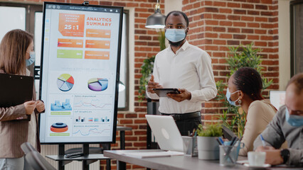 Man and woman with face mask planning business growth in front of coworkers at briefing meeting. Colleagues using monitor to do financial presentation for marketing strategy during pandemic.