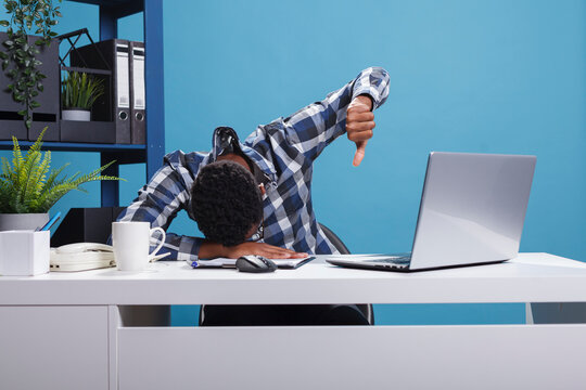 Distressed and exhaused hard working company employee falling asleep on desk while showing disapproval symbol with hand. Sleepy tired man showing dislike gesture while resting head on desk.