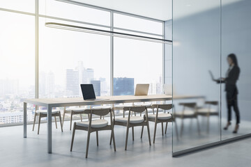 Attractive young european businesswoman with laptop standing in modern concrete office interior with matte partition glass, furniture, equipment and city view with daylight. Worker and ceo concept.