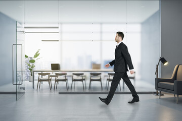 Attractive young european businessman walking in modern concrete office interior with matte partition glass, furniture, equipment and city view with daylight. Worker and ceo concept.
