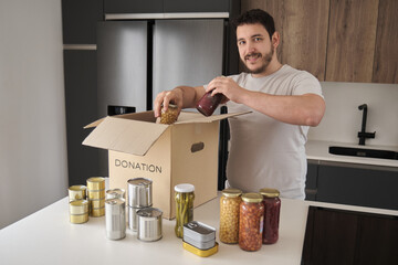 Young latin man smiling and filling donation box with food cans, legume jars and canned fish in...
