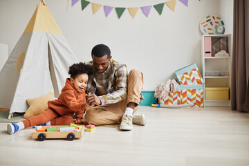 Full length portrait of caring black father and son playing with toys together in minimal kids room...