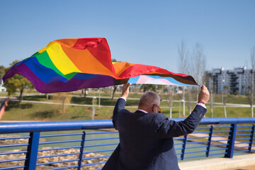 Mature gay man, executive, gray haired, with beard, sunglasses and jacket, walking with new lgbtiq+ pride flag in the wind above his head. Concept mature gay man, suggar daddy, lgbt, pride, teddy bear