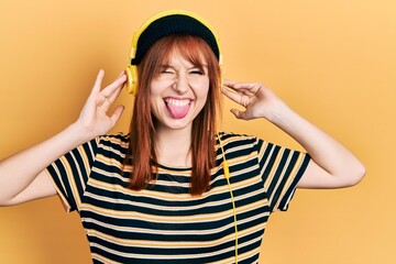 Obraz na płótnie Canvas Redhead young woman listening to music using headphones sticking tongue out happy with funny expression.
