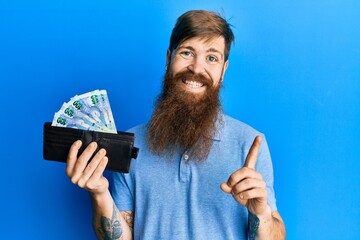Redhead man with long beard holding wallet with south african rand banknotes smiling happy pointing...