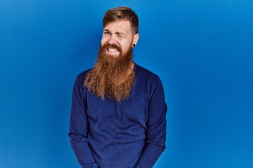 Redhead man with long beard wearing casual blue sweater over blue background winking looking at the camera with sexy expression, cheerful and happy face.