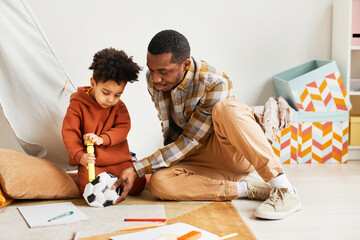 Full length portrait of caring black father playing with toddler son indoors and pumping up kids soccer ball, copy space
