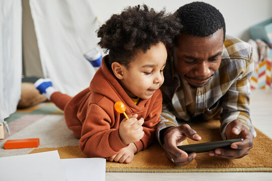 Portrait of black father with toddler boy laying on floor together and watching cartoons via smartphone in playroom