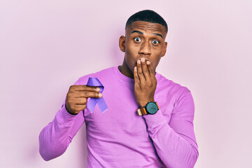 Young black man holding purple ribbon awareness covering mouth with hand, shocked and afraid for...