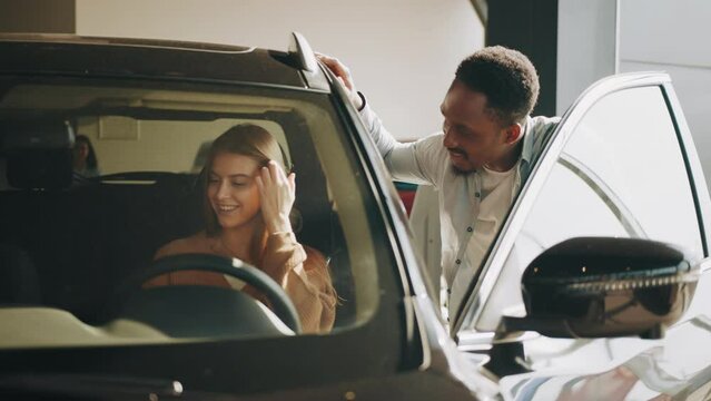 Cheerful caucasian woman sitting inside luxury car and testing leather interior while african american delaer standing near and providing competent consultation. Concept auto business. Dealer car.
