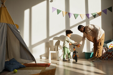 Minimal portrait of loving black father teaching little boy riding balance bike at home in sunlight, copy space