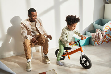 Full length portrait of caring black father teaching little boy riding balance bike at home in...