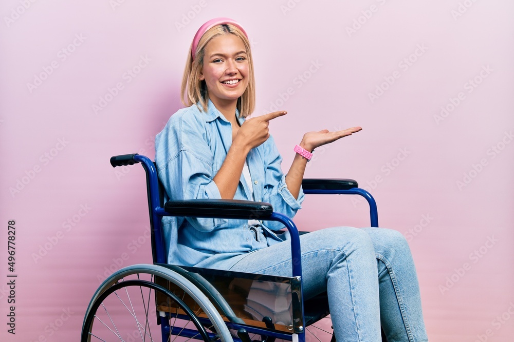 Sticker Beautiful blonde woman sitting on wheelchair amazed and smiling to the camera while presenting with hand and pointing with finger.