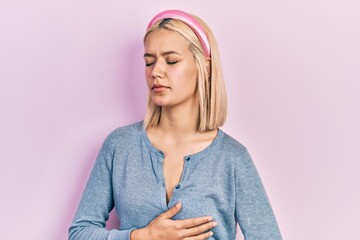 Beautiful blonde woman standing over pink background with hand on stomach because indigestion, painful illness feeling unwell. ache concept.