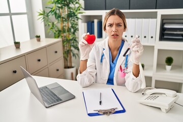 Young blonde woman wearing doctor uniform holding blood and heart clueless and confused expression....