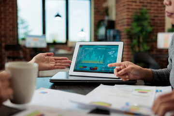 Clsoe up of digital tablet with management graphs on display standing on table in startup office during business meeting. Businesswomen discussing company strategy working at partnership project
