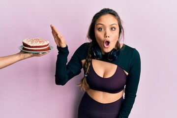 Young hispanic girl wearing sportswear rejecting cake scared and amazed with open mouth for...