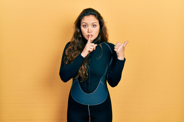 Young hispanic girl wearing diver neoprene uniform asking to be quiet with finger on lips pointing...