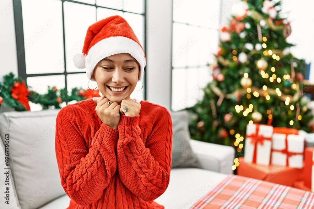 Wall mural Young hispanic woman with short hair wearing christmas hat sitting on the sofa laughing nervous and excited with hands on chin looking to the side