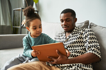 Portrait of young black father and little boy using digital tablet together while relaxing on couch at home