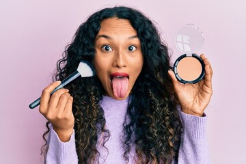 Young latin woman holding makeup brush and blush sticking tongue out happy with funny expression. © Krakenimages.com