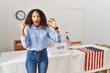 Beautiful hispanic woman standing by at political campaign by voting ballot surprised pointing with hand finger to the side, open mouth amazed expression.