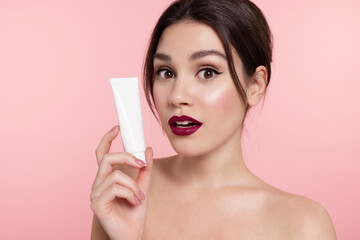 Surprised beautiful young woman show mockup cosmetics tube with lotion, cream or makeup foundation. Modern girl with trendy make up holding cosmetic package, advertise natural skincare beauty product.