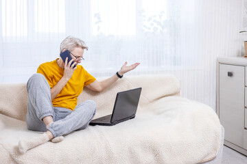 adult woman is emotionally talking on the phone while sitting at a laptop on the couch at home