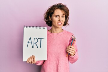 Young brunette woman holding art notebook and colored pencils clueless and confused expression. doubt concept.