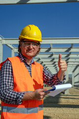 Construction Worker With Clipboard Giving Thumbs Up At Construction Site. Construction Foreman Holding Clipboard and Looking at Camera. Worker in Yellow Hardhat  Standing at Construction Site.