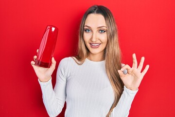 Naklejka premium Young blonde girl holding shampoo bottle doing ok sign with fingers, smiling friendly gesturing excellent symbol