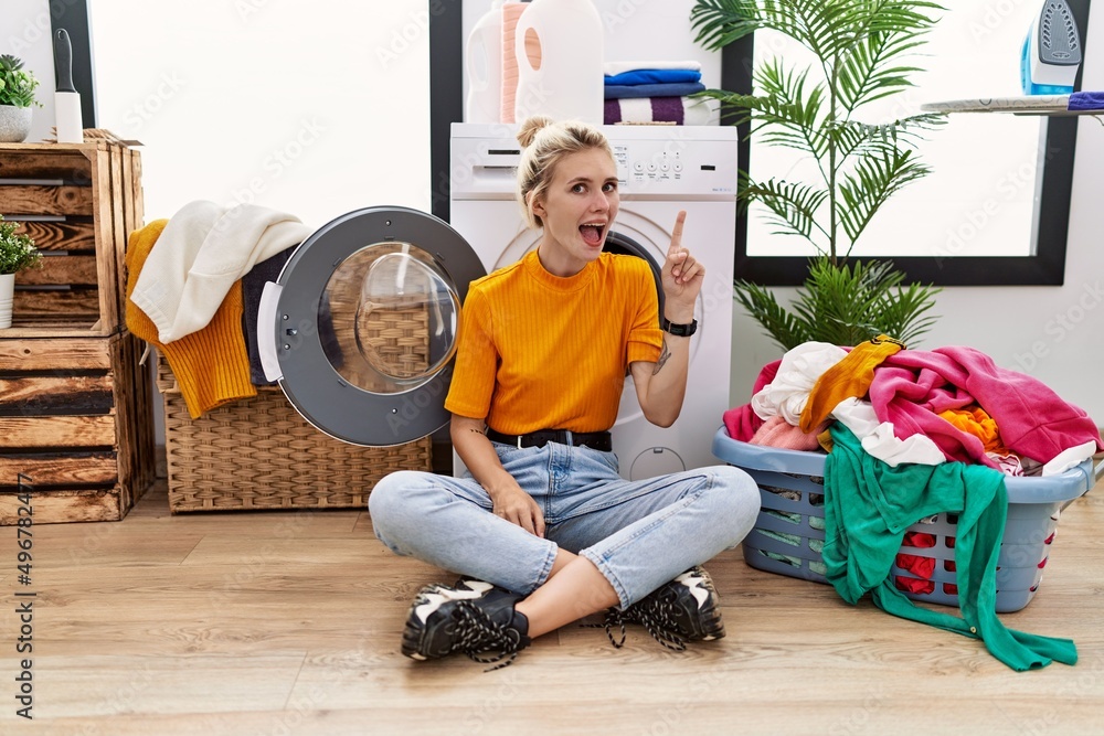 Poster young blonde woman doing laundry sitting by washing machine pointing finger up with successful idea.