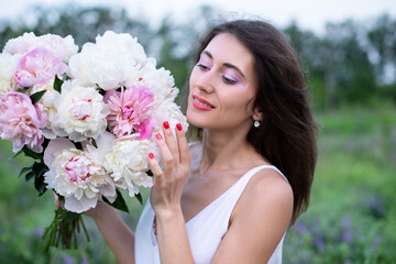 Smiling woman with a bouquet of peonies. Close-up portrait of a beautiful brunette woman among peony flowers. Beauty and flowers. Cosmetics, makeup. Perfumery. Closeup of charming lady with flowers	
