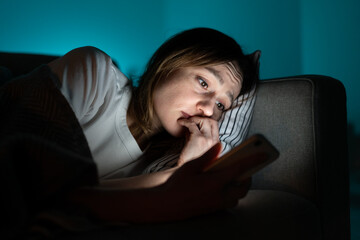 Young anxious woman lying in bed staring at smartphone screen at night, reading about depression symptoms in internet, phone addicted female can not stop scrolling news media before bedtime. Anxiety. - obrazy, fototapety, plakaty