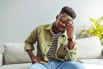 Young African man touching head with hands and keeping eyes closed while sitting on the sofa at home. Black guy stressing and headache. Feeling sick and tired.