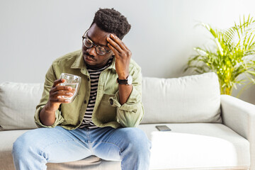 Young man suffering from strong headache or migraine sitting with glass of water in the living room, millennial guy feeling intoxication and pain touching aching head, morning after hangover concept