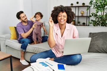 Mother of interracial family working using computer laptop at home gesturing finger crossed smiling with hope and eyes closed. luck and superstitious concept.