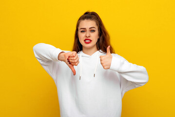 Pretty brunette girl in white casual style sweatshirt is showing thumbs up and thumbs down, disagreement and agreement expression, concept of difficult choice. Indoor studio shot on yellow background - obrazy, fototapety, plakaty