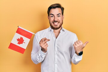 Handsome caucasian man with beard holding canada flag pointing thumb up to the side smiling happy with open mouth