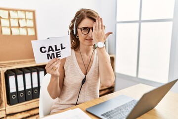 Middle age brunette woman wearing operator headset holding call me banner covering one eye with...