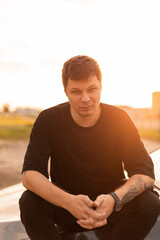 Serious business cool man hipster with a black T-shirt sits on the hood of a car at sunset. Summer menswear style