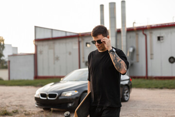 Cool stylish hipster man with a longboard and a black T-shirt is fixing his sunglasses and walking near a car on the street