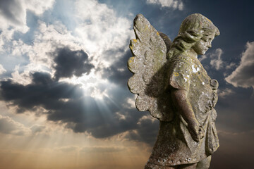 Stone statue of winged angel against rays of sunlight behind clouds