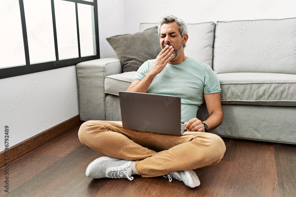 Sticker middle age hispanic man using laptop sitting on the floor at the living room bored yawning tired cov