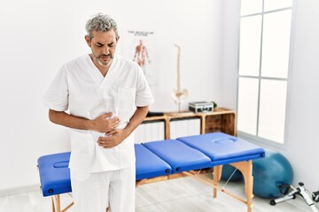 Middle age hispanic therapist man working at pain recovery clinic with hand on stomach because indigestion, painful illness feeling unwell. ache concept.
