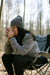 Fototapeta premium Young woman sitting on camp chair. She is drinking hot drinks. Coffee or tea. She has hat. Forest background.