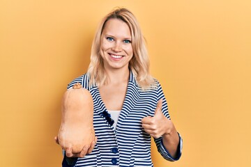 Beautiful caucasian blonde woman holding healthy fresh pumpkin smiling happy and positive, thumb up doing excellent and approval sign