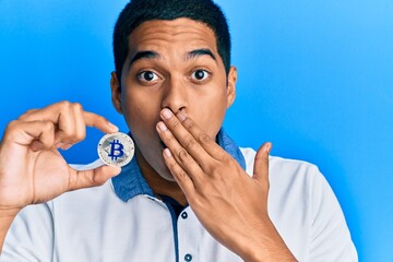 Young handsome hispanic man holding virtual currency bitcoin covering mouth with hand, shocked and...