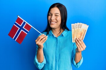 Young latin woman holding norway flag and krone banknotes winking looking at the camera with sexy...